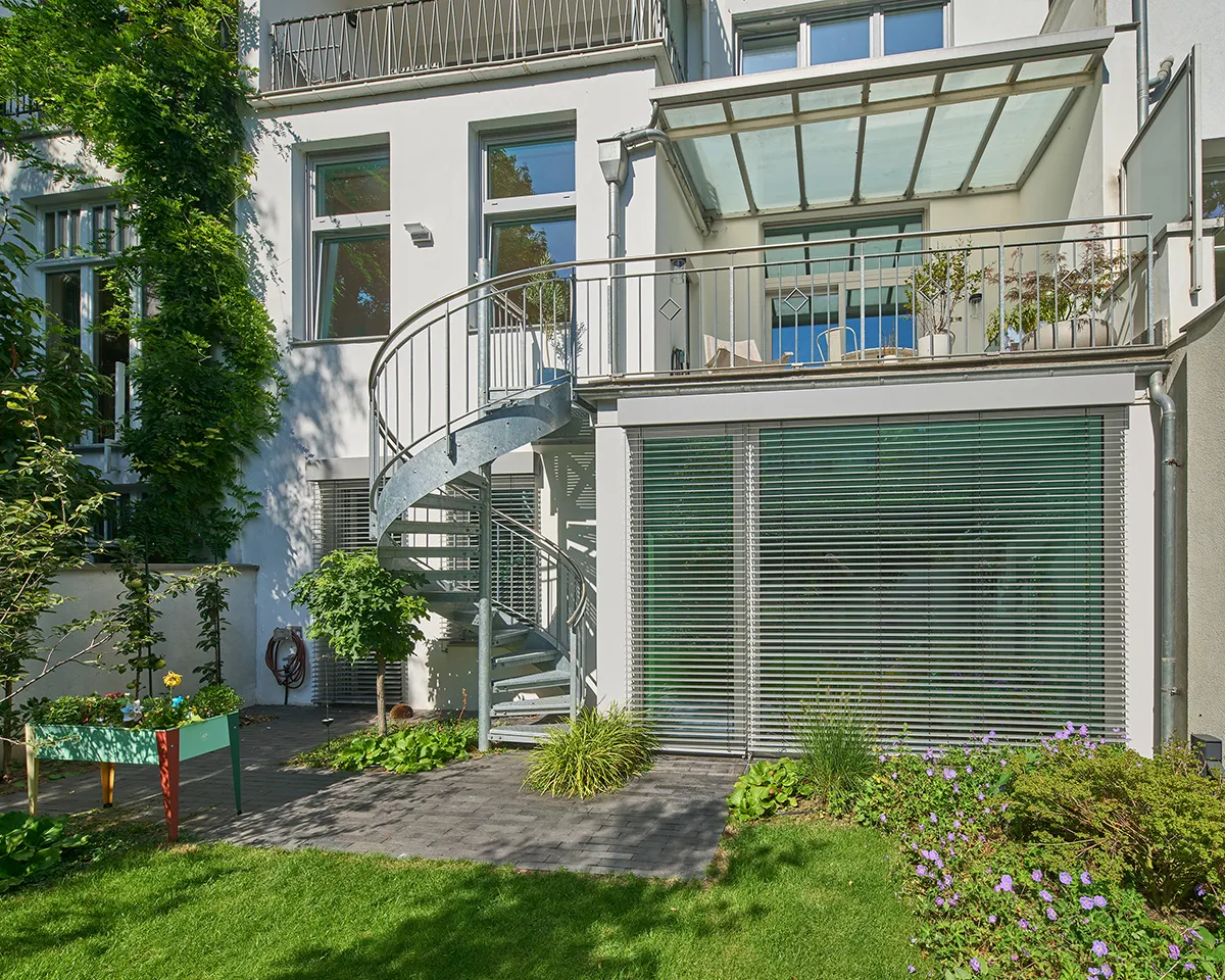 Moderne Gartenansicht einer umgebauten Maisonettewohnung mit verglaster Terrasse, Wendeltreppe aus Metall und gepflegtem Gartenbereich.